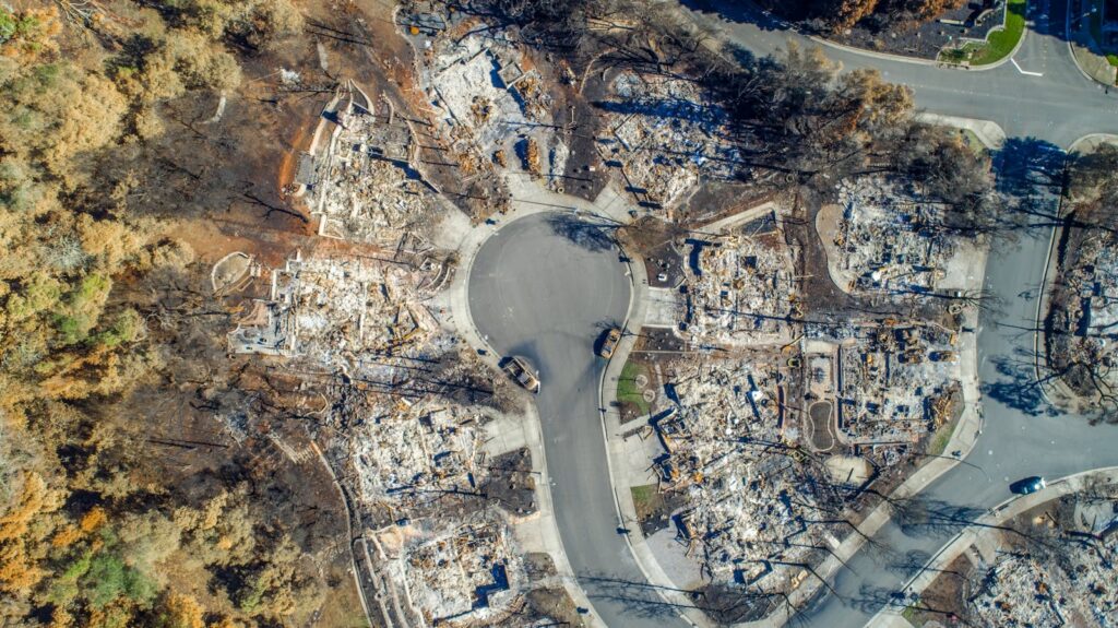 Aerial view of a residential cul‑de‑sac devastated by wildfire, with homes reduced to charred foundations, burned trees surrounding the area, and scattered debris visible along the circular street.